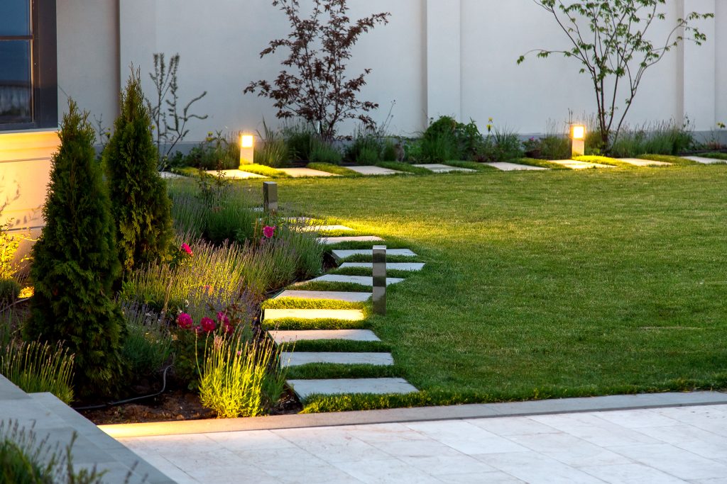 Backyard marble walkway and flowerbed lit by decorative garden lighting at dusk with warm ground lamps.
