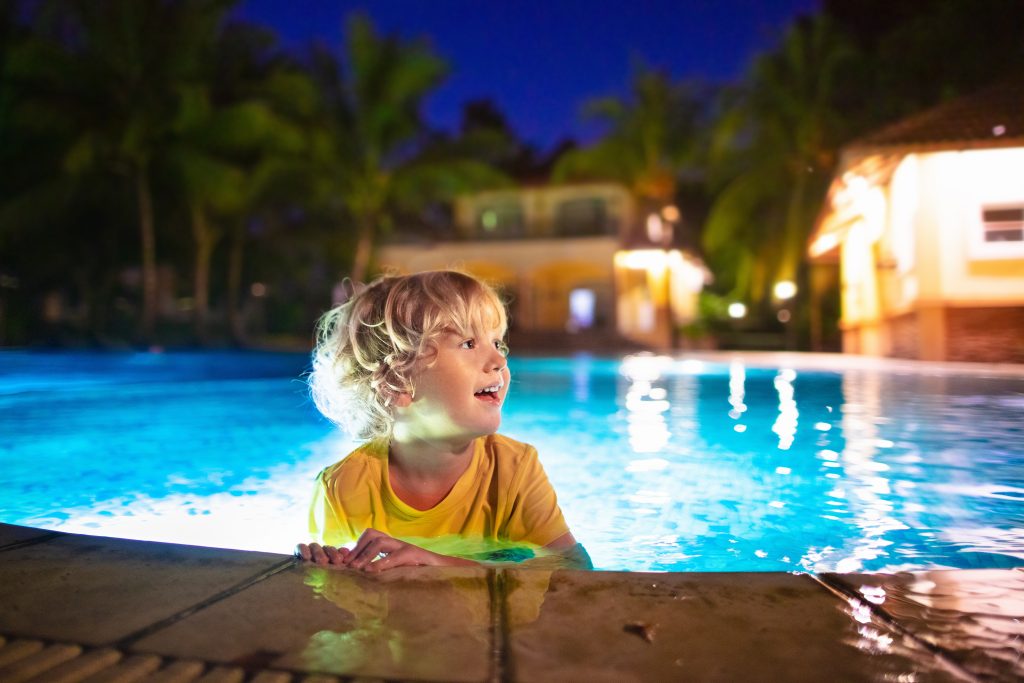 Child smiling at the edge of a well-lit backyard swimming pool at night with home visible behind.